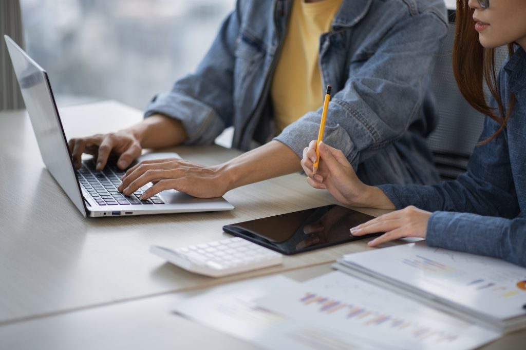Mãos de pessoas trabalhando juntas em mesa com computador e cadernos, representando colaboração e atividades práticas em curso técnico.